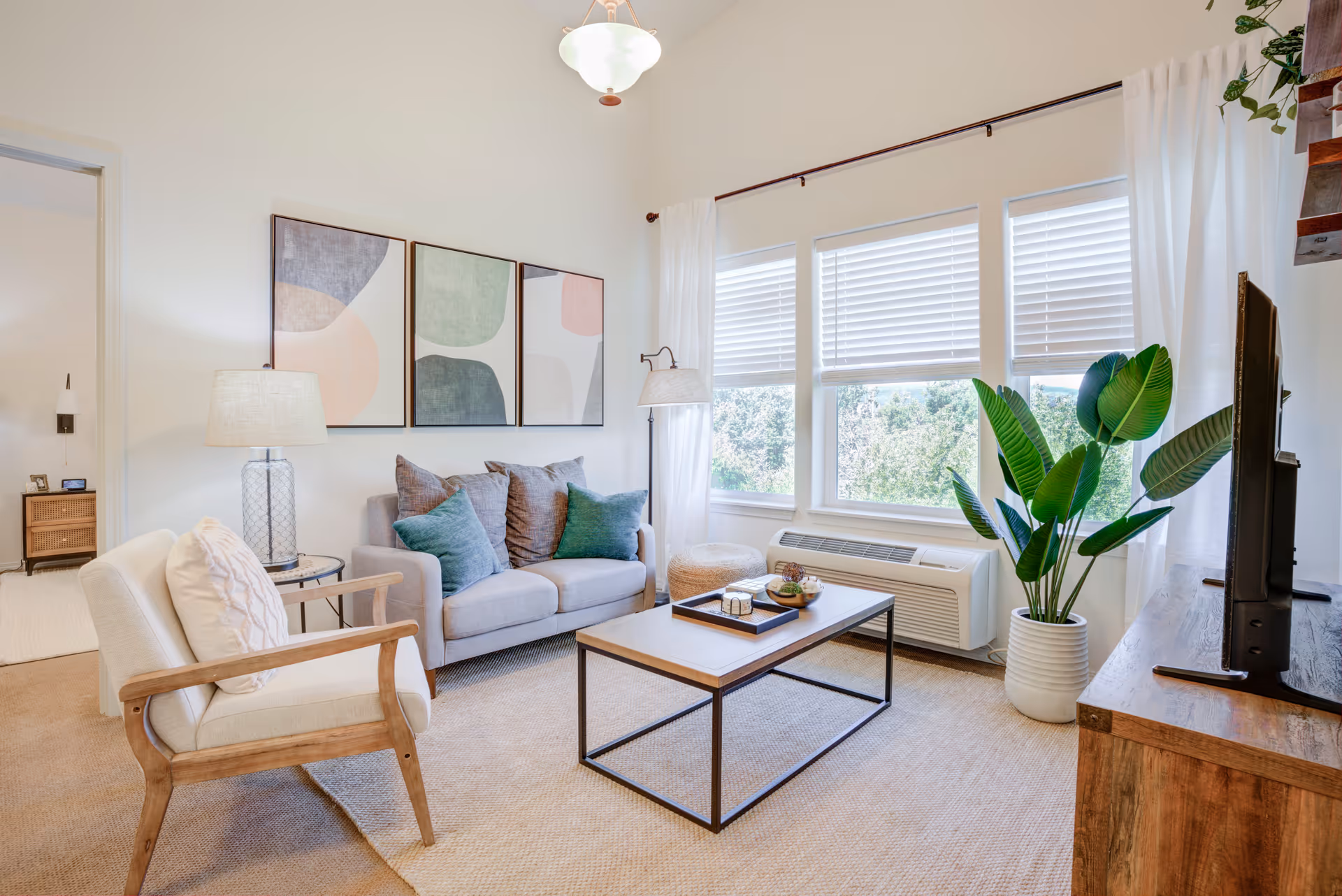 Bright furnished living room with a sofa, armchair, coffee table, TV stand, and large windows with white blinds.