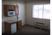 A small room with a wooden cabinet set including a microwave on the upper shelf and a mini refrigerator below. There is a closed white door next to the cabinet. A window with blinds is on the adjacent wall, and below it is a wall-mounted air conditioning unit. The floor is carpeted.
