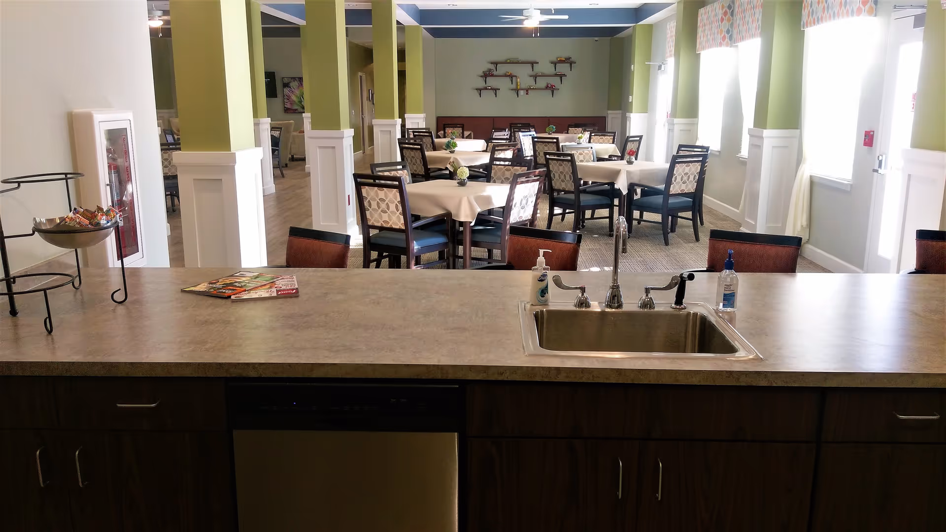View from a kitchen counter with a sink and hand sanitizer bottles looking out into a dining area with multiple tables and chairs, green walls, and large windows letting in natural light.