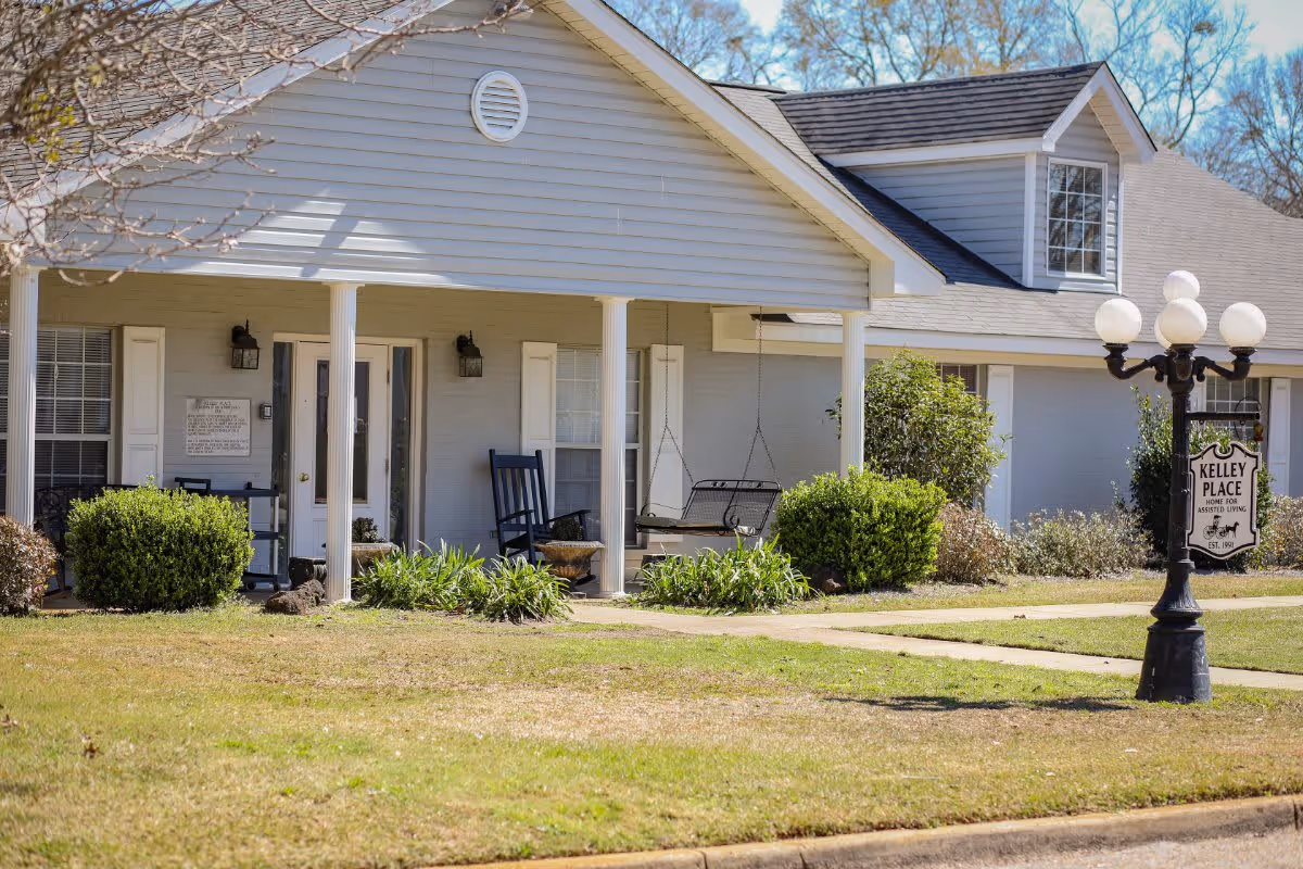Exterior view of Kelley Place, a home for assisted living, showing a single-story building with a covered porch. The porch has a rocking chair and a swing, surrounded by bushes and plants. A vintage-style street lamp with a sign that reads 'Kelley Place Home for Assisted Living Est. 1991' is visible on the lawn.