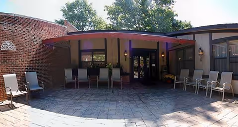Outdoor patio area with several chairs arranged along the walls and under a red awning attached to a building. The building has brick and siding exterior walls, with windows and outdoor wall lanterns. Trees are visible in the background.