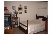 A small bedroom with a single bed covered with a beige bedspread. There is a wooden dresser on the right side of the bed and a bookshelf filled with books and decorative items on the left. A television sits on a wooden stand in the corner. Several framed pictures and a clock hang on the wall above the bookshelf. The room has a simple, cozy appearance.