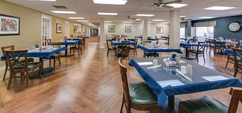 A spacious dining room with multiple tables covered with blue tablecloths, each set with placemats, napkins, and utensils. Wooden chairs with green cushions surround the tables. The room has wood-patterned flooring, ceiling fans, framed artwork on the walls, and large windows letting in natural light.