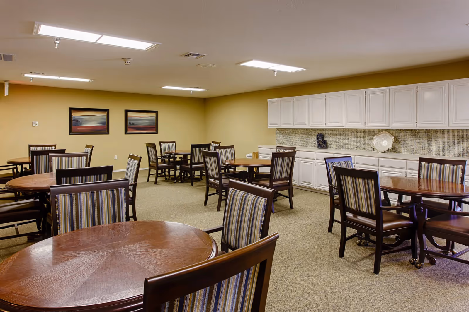 A senior living facility dining room with multiple round wooden tables and striped cushioned chairs. The room has beige walls, carpeted floor, white cabinets along one wall, and two framed landscape paintings on the far wall. The ceiling has recessed lighting panels.
