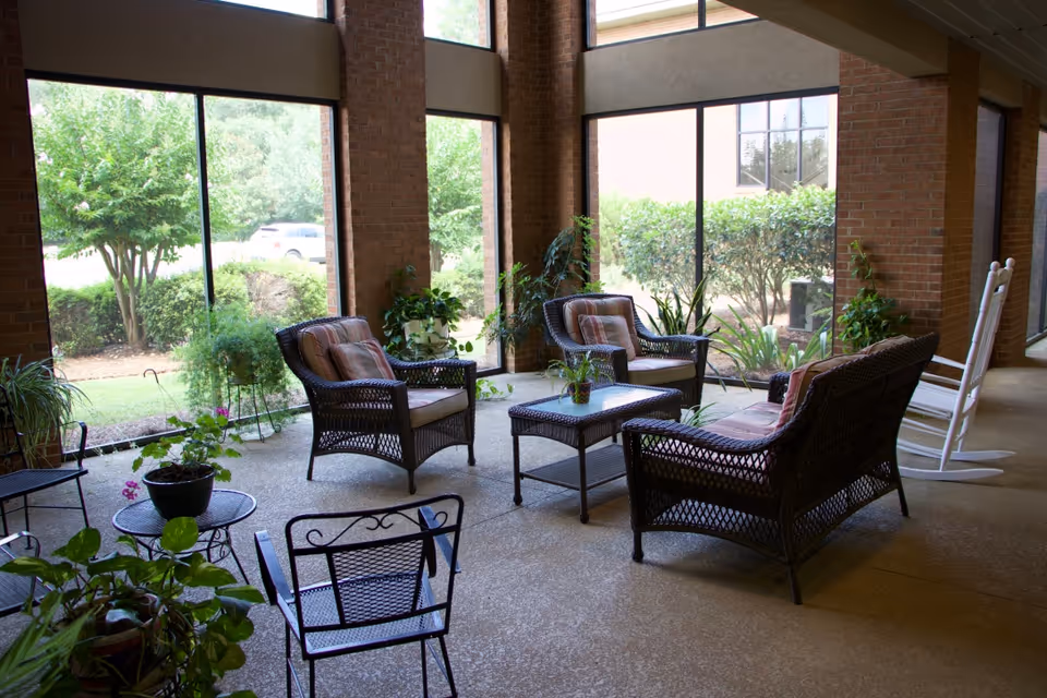 Sunlit indoor seating area with wicker chairs, a coffee table, plants, and rocking chairs by large windows overlooking greenery.