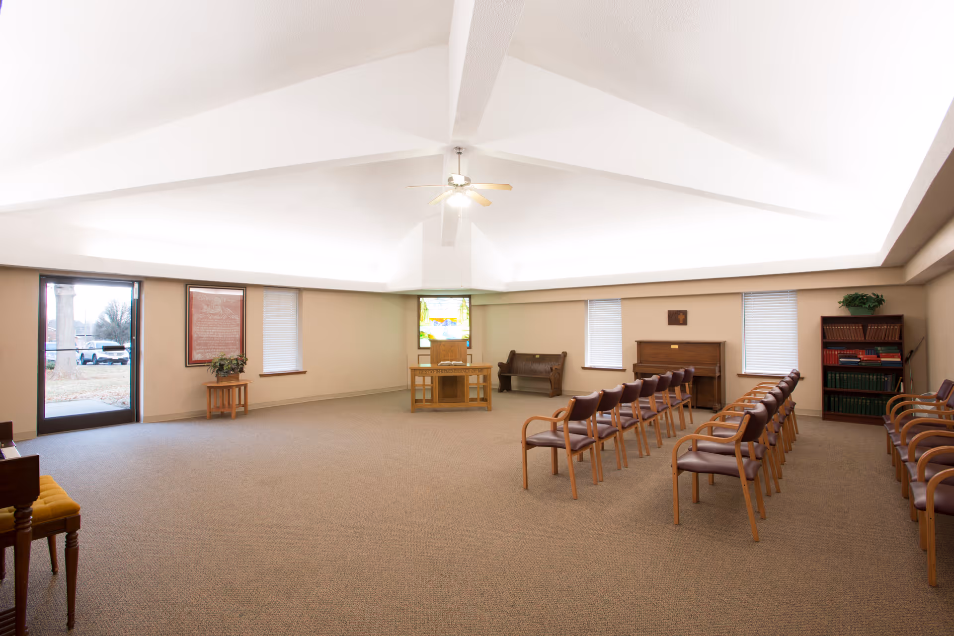 A spacious room with beige walls and carpeted floor, featuring rows of wooden chairs with brown cushions arranged facing a small wooden podium with a stained glass window behind it. The room has a high, white ceiling with a ceiling fan in the center. There is a piano against the far wall, a wooden bench, a bookshelf with books, and a door with windows letting in natural light.