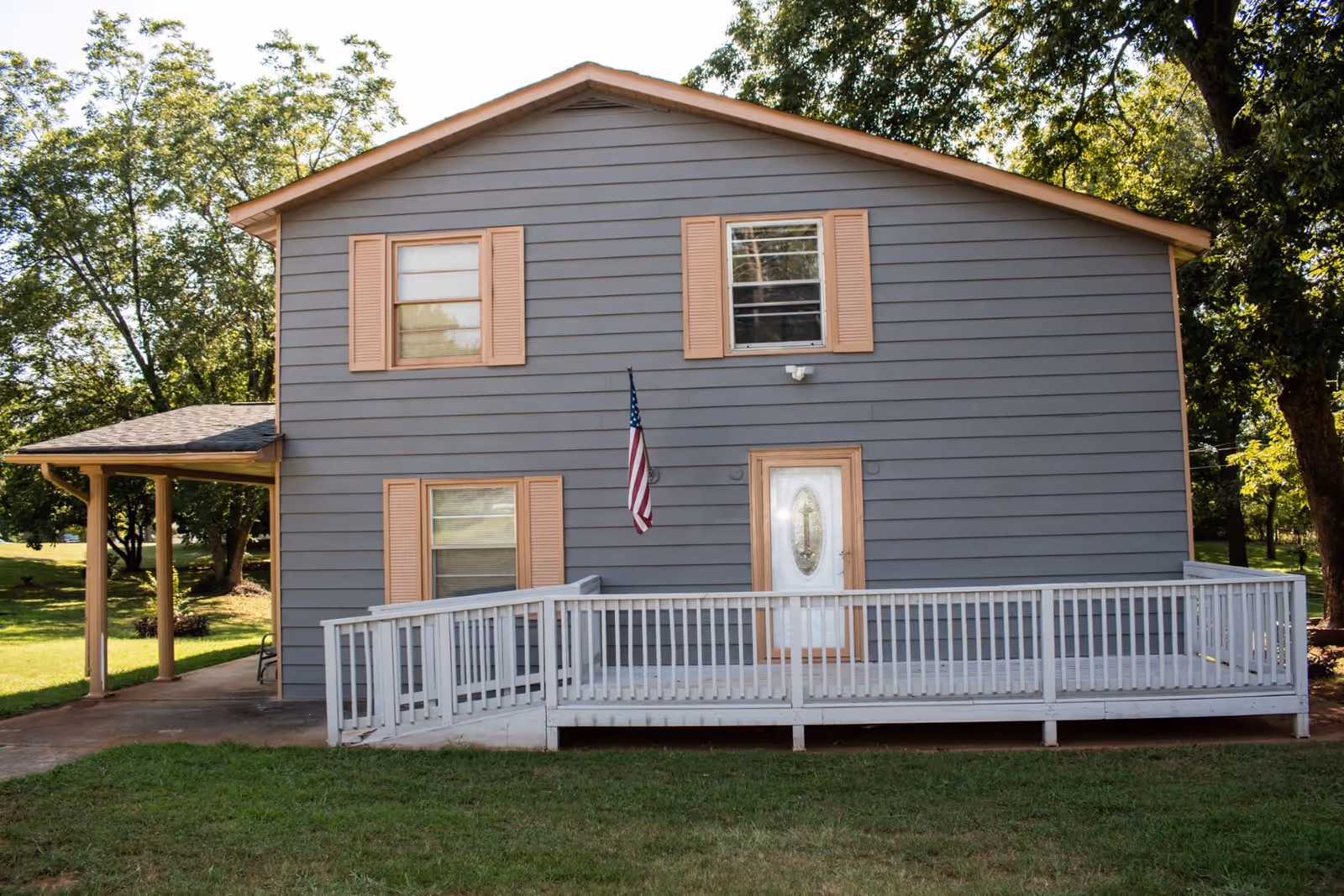Exterior view of a two-story gray house with peach-colored window shutters and a white front door with decorative glass. There is a white wooden ramp leading to the front door and an American flag mounted on the wall. The house is surrounded by green grass and trees.