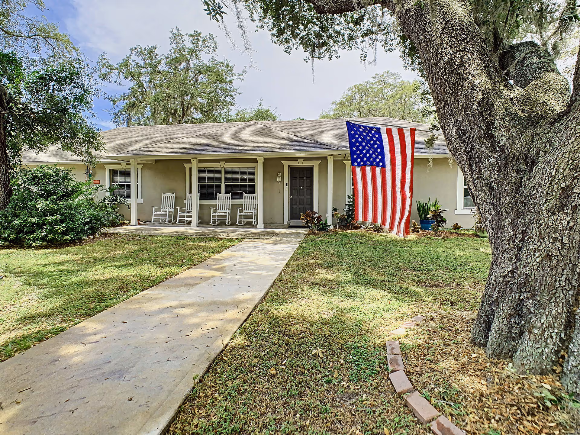 Front exterior view of a single-story building with a covered porch featuring four white rocking chairs. An American flag is hanging near the entrance. There is a concrete walkway leading up to the front door, surrounded by grass and trees.