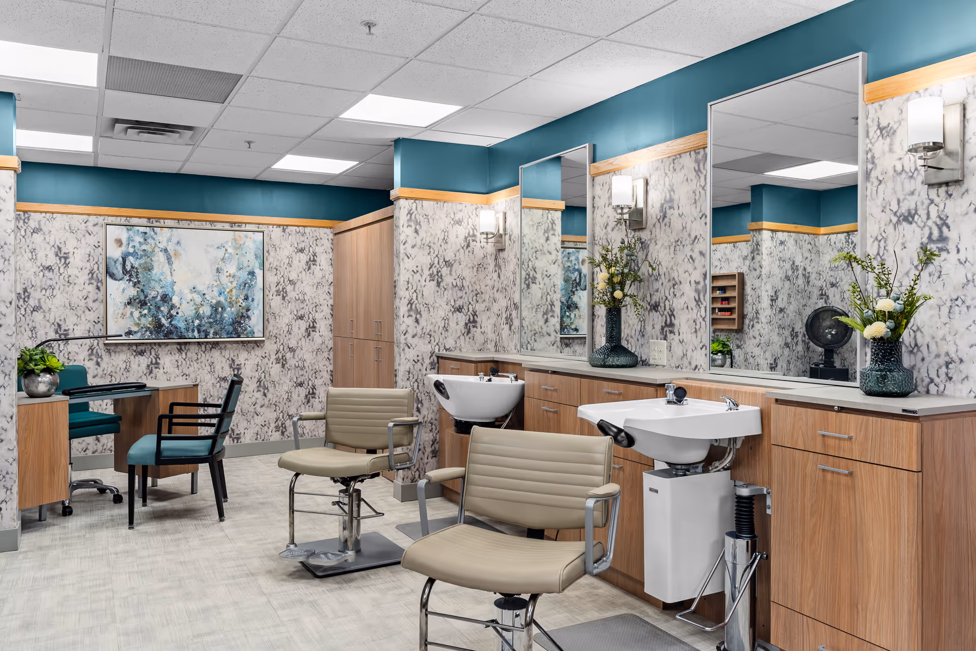 Interior view of a salon area in a senior living facility with two beige salon chairs in front of sinks and large mirrors. The walls have a marble-like pattern with teal accents and wooden trim. There is a small desk with two teal chairs and a decorative painting on the wall.