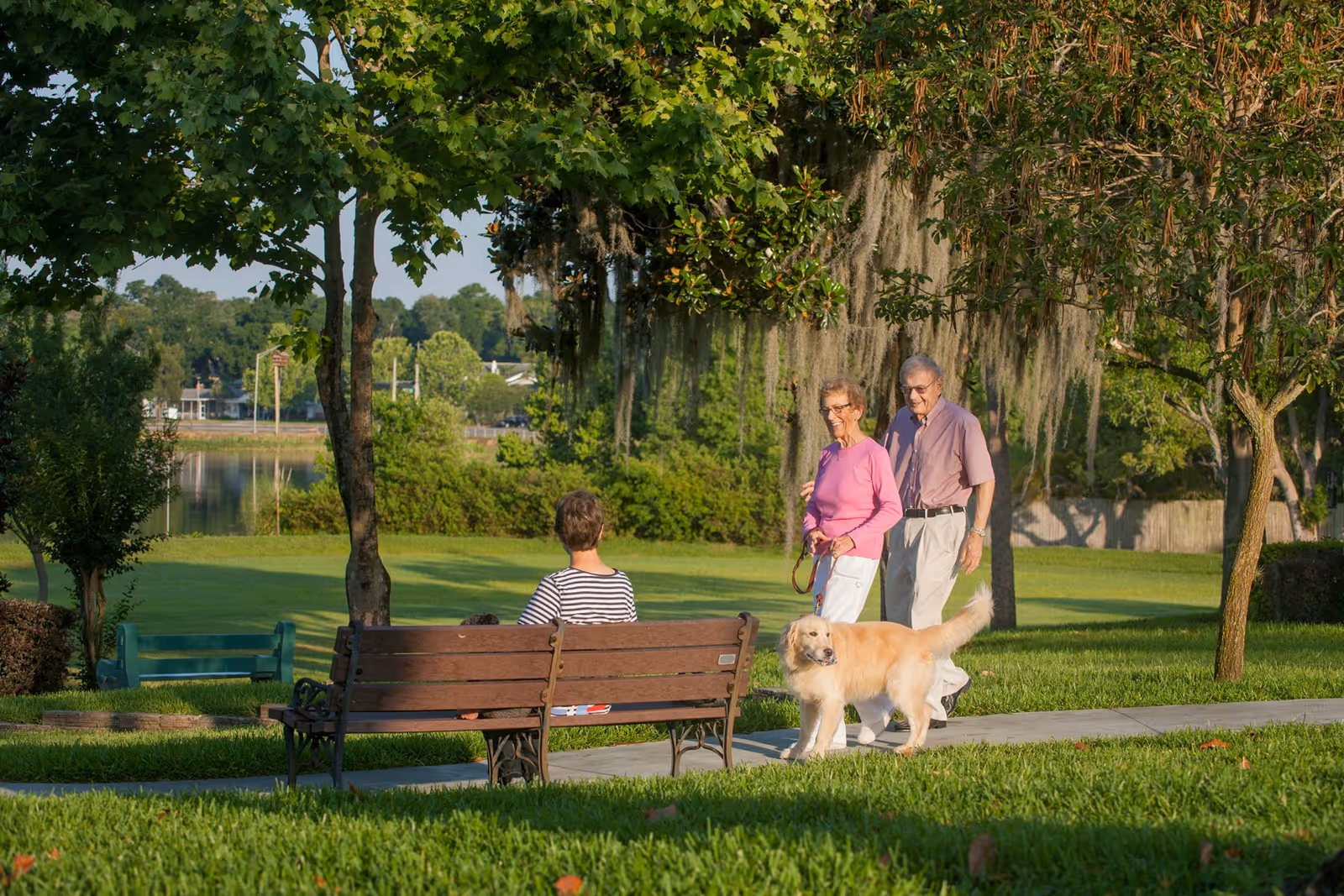 A couple walks a golden retriever on a paved path beside a bench where a person sits in a leafy park by a lake.
