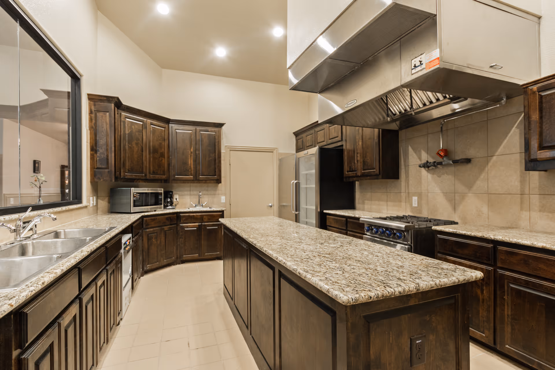 A spacious kitchen with dark wooden cabinets, granite countertops, a large central island, a stainless steel stove with an overhead exhaust hood, a microwave, a coffee maker, and a double sink. The kitchen has beige tiled walls and floor, and recessed ceiling lights.