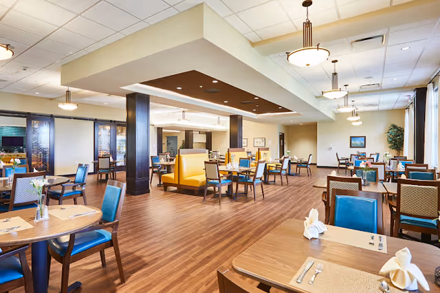 Spacious dining room with wooden floors, multiple tables and chairs arranged neatly, yellow booth seating in the center, pendant lights hanging from the ceiling, and large windows with curtains allowing natural light to fill the room.