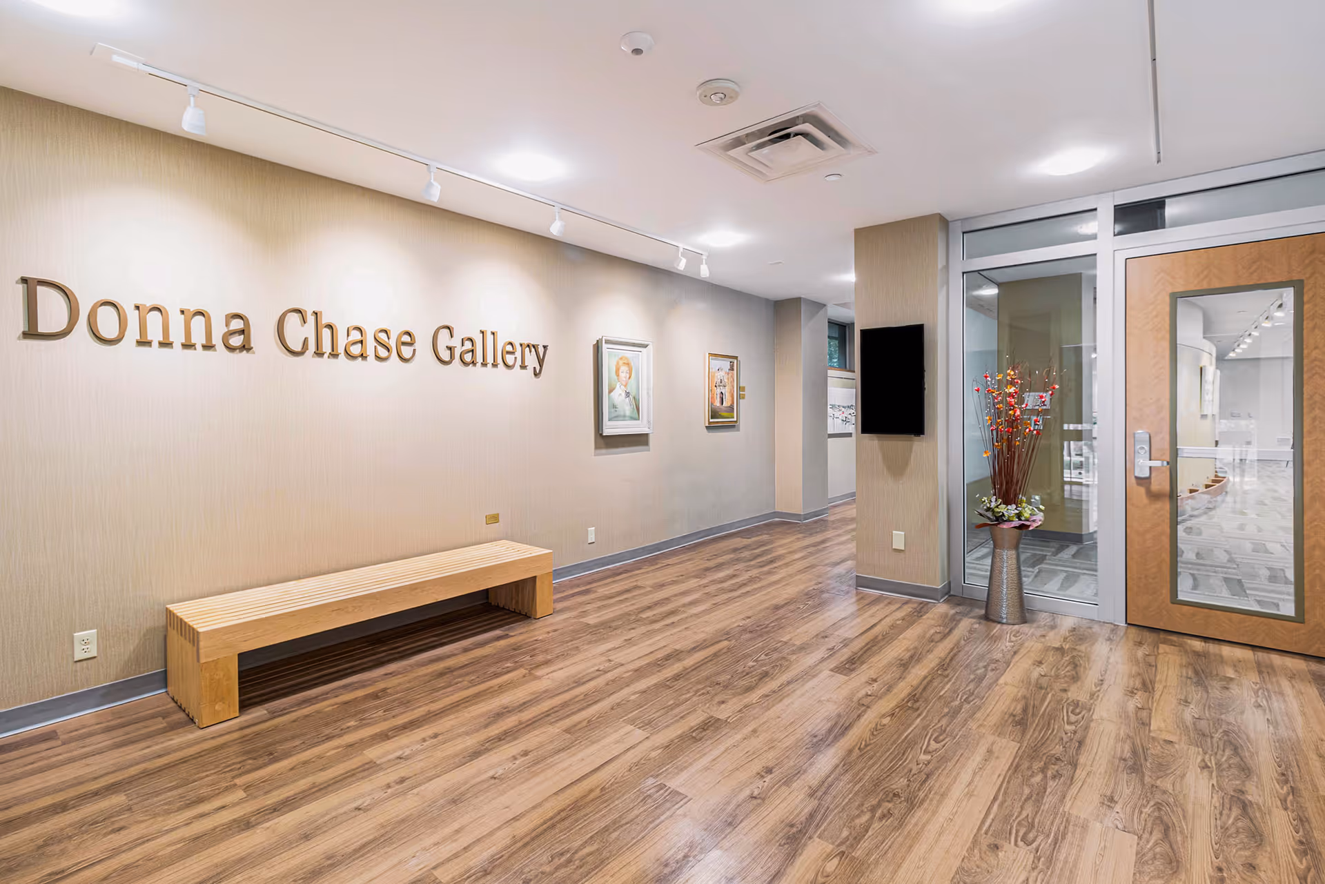 Interior gallery foyer labeled 'Donna Chase Gallery' with a wooden bench, framed paintings, a vase with flowers, and a glass door.