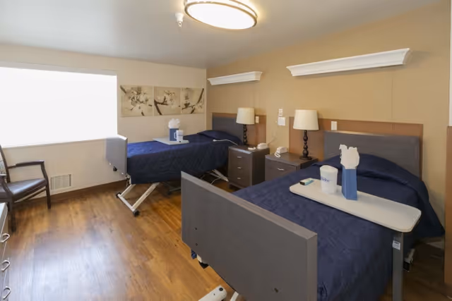 A room with two hospital-style beds covered with dark blue quilts, each with a tray holding a tissue box and a cup. Between the beds is a nightstand with two lamps, a telephone, and a remote control. There is a window with a white blind on the left wall and a wooden chair next to it. The floor is wooden, and the walls are painted beige with white crown molding accents.