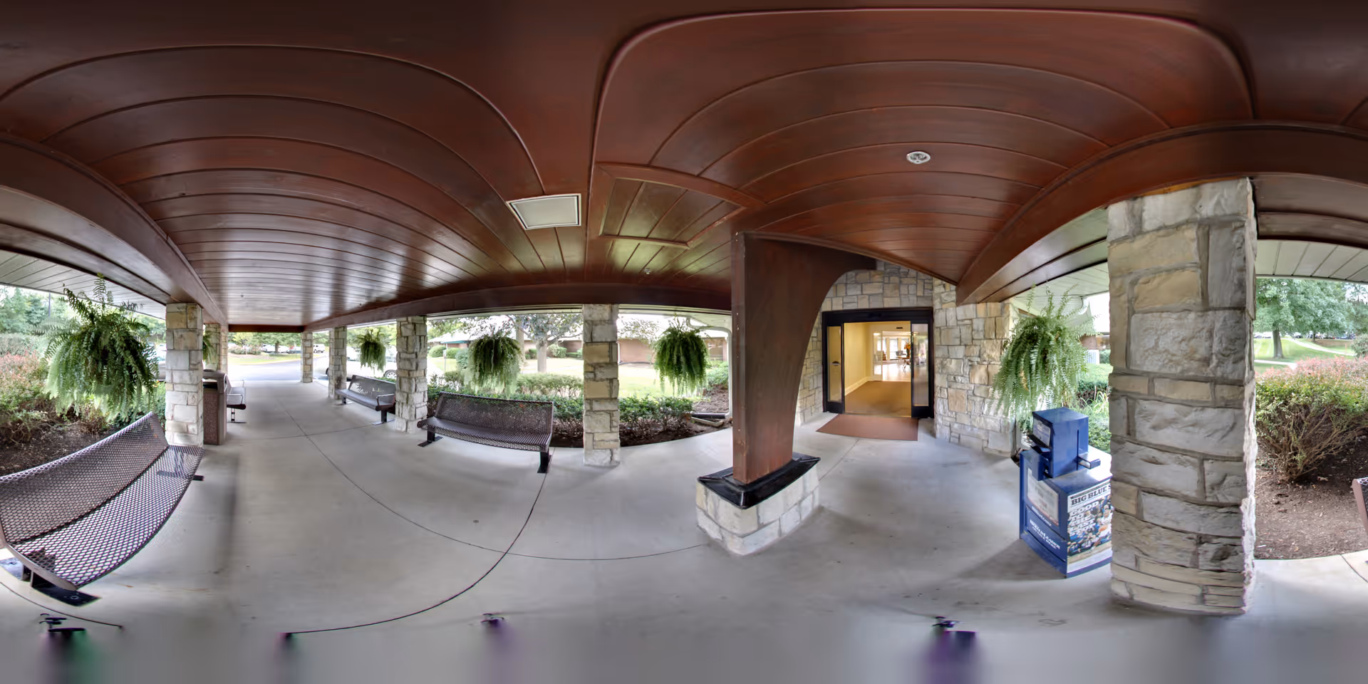 Covered outdoor walkway with stone pillars and wooden ceiling at the entrance of a building. There are hanging ferns, metal benches along the sides, and a newspaper vending machine near the entrance door. Green bushes and trees are visible outside the walkway.