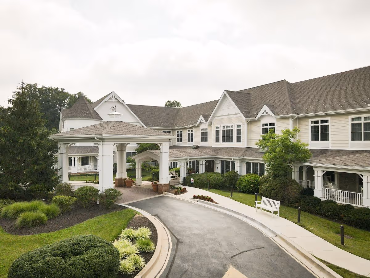 Front exterior of a two-story senior living facility with a covered porte-cochere, driveway, benches, and landscaped grounds.