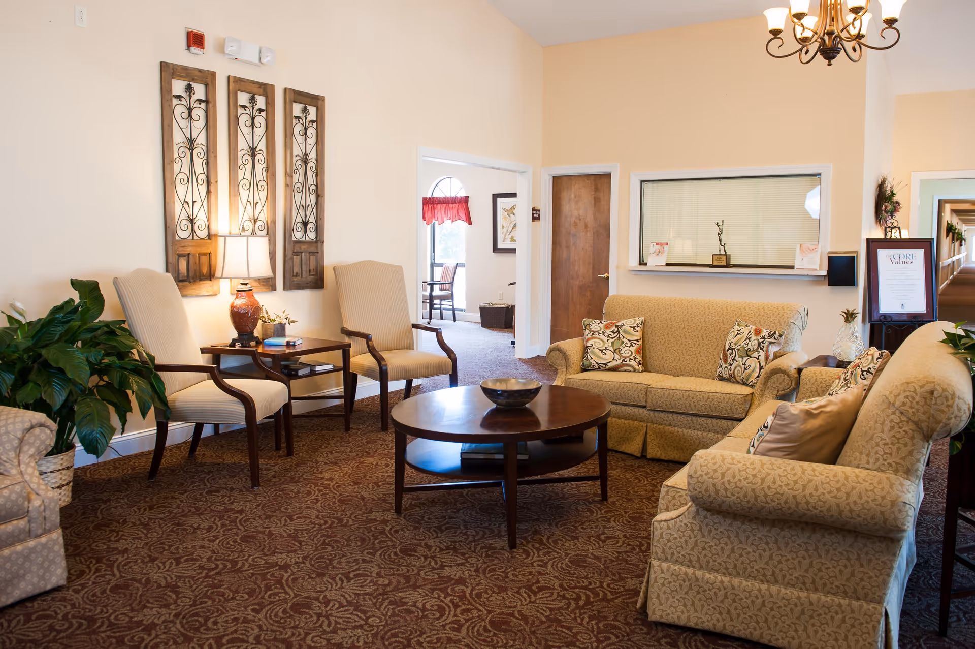 A cozy senior living facility common area with beige upholstered armchairs and sofas arranged around a round wooden coffee table. The room features a patterned carpet, a table lamp on a side table, decorative wall art, and a chandelier hanging from the ceiling. There are plants placed around the room and a hallway visible in the background.
