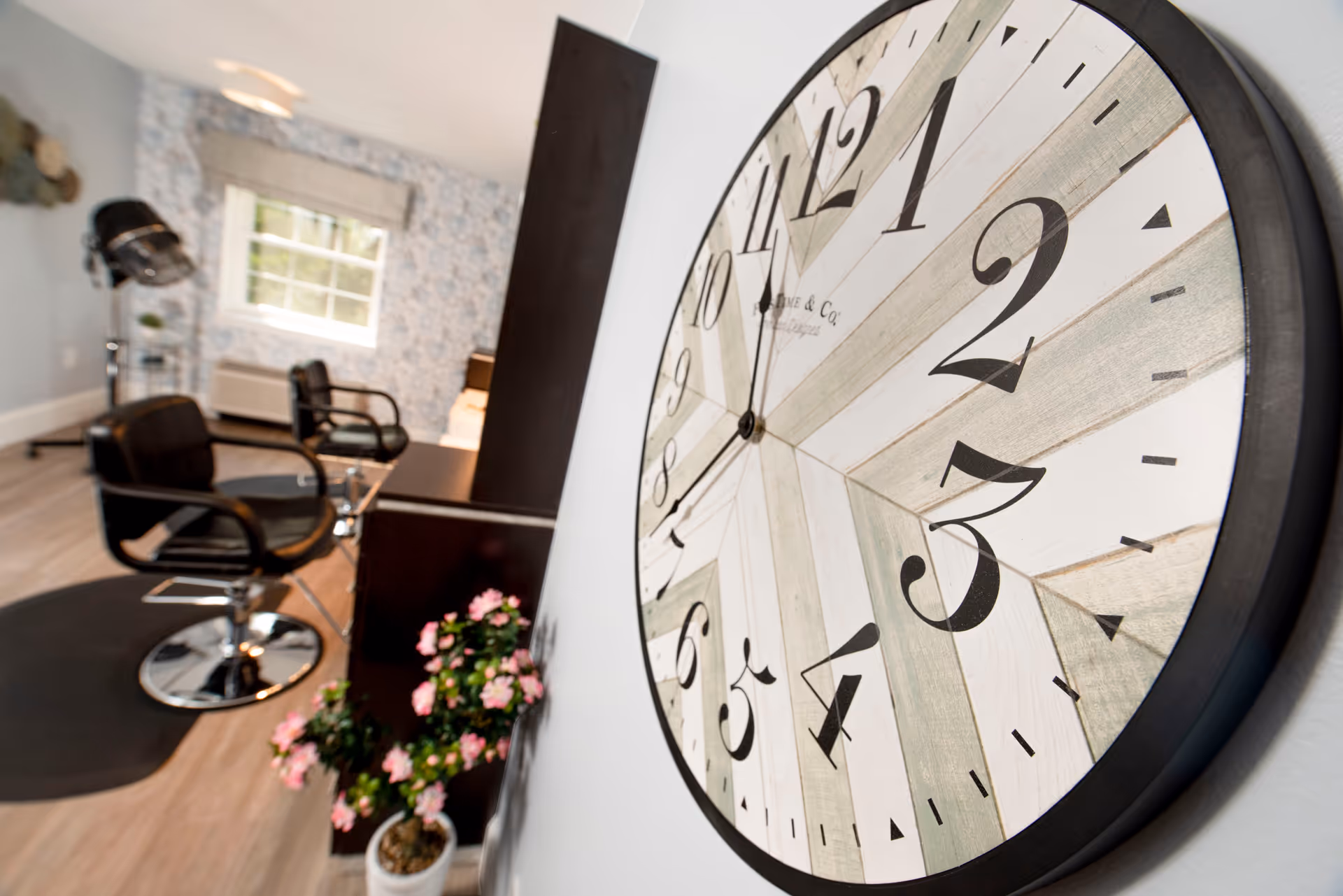 Close-up of a large decorative wall clock with salon styling chairs, a hood dryer and potted flowers visible in the background of an interior room.