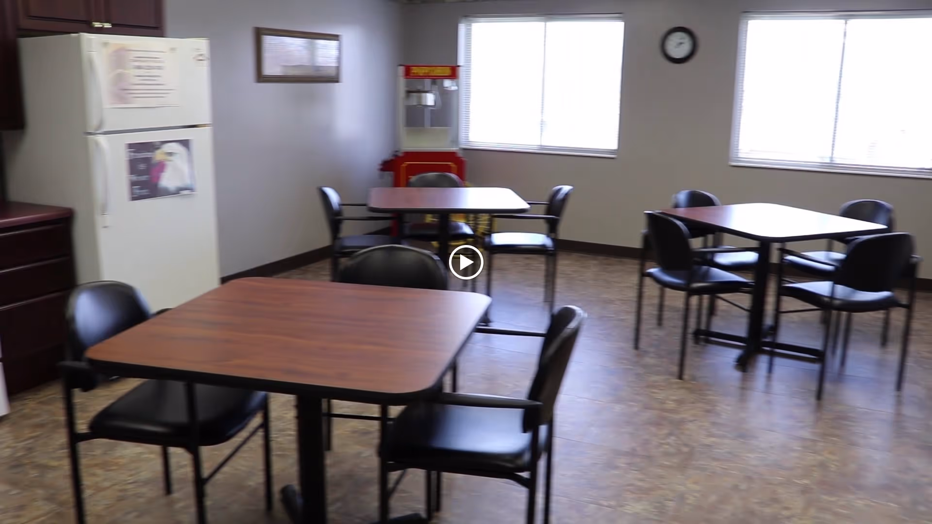 A dining area with three square wooden tables, each surrounded by four black chairs. There is a white refrigerator on the left side, a popcorn machine near the back wall, two large windows letting in natural light, and a clock on the wall between the windows.