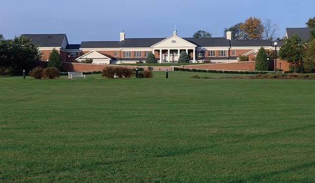 Wide view of a low red-brick senior living building with a columned central entrance and a large green lawn in the foreground.