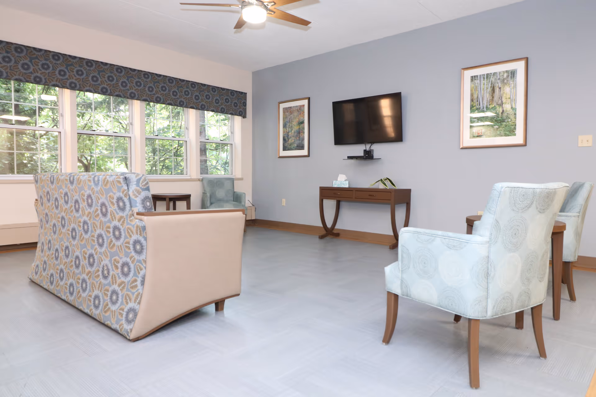 Bright common living room with patterned armchairs and a sofa facing a wall-mounted TV and large windows.