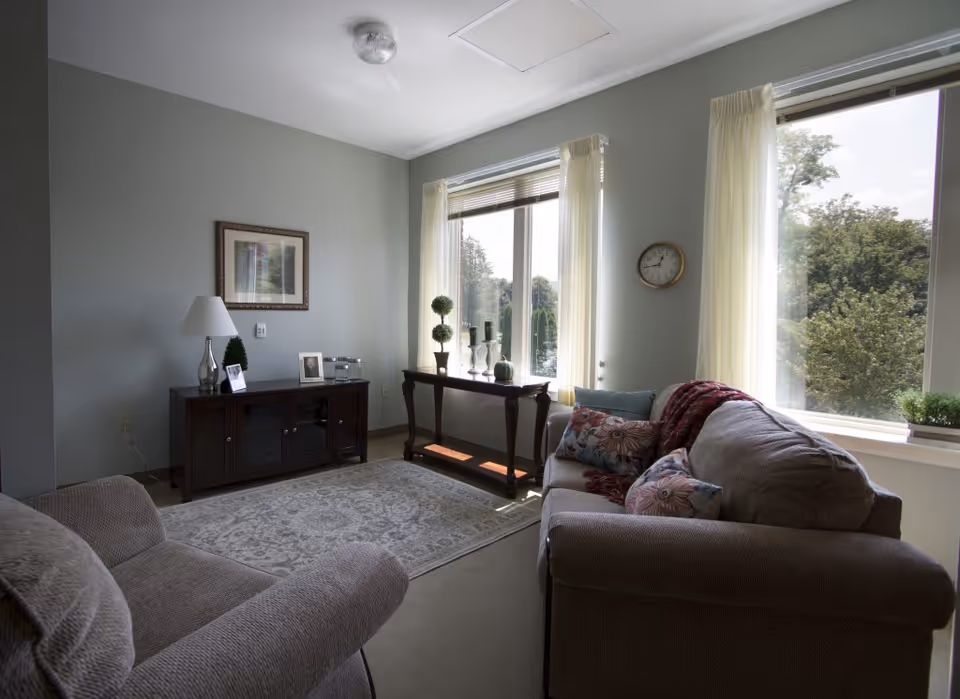A cozy living room with a beige sofa and armchair, floral patterned pillows, a red throw blanket, a wooden console table with decorative items, a framed picture on the wall, a table lamp, and large windows with sheer curtains letting in natural light and showing green trees outside.