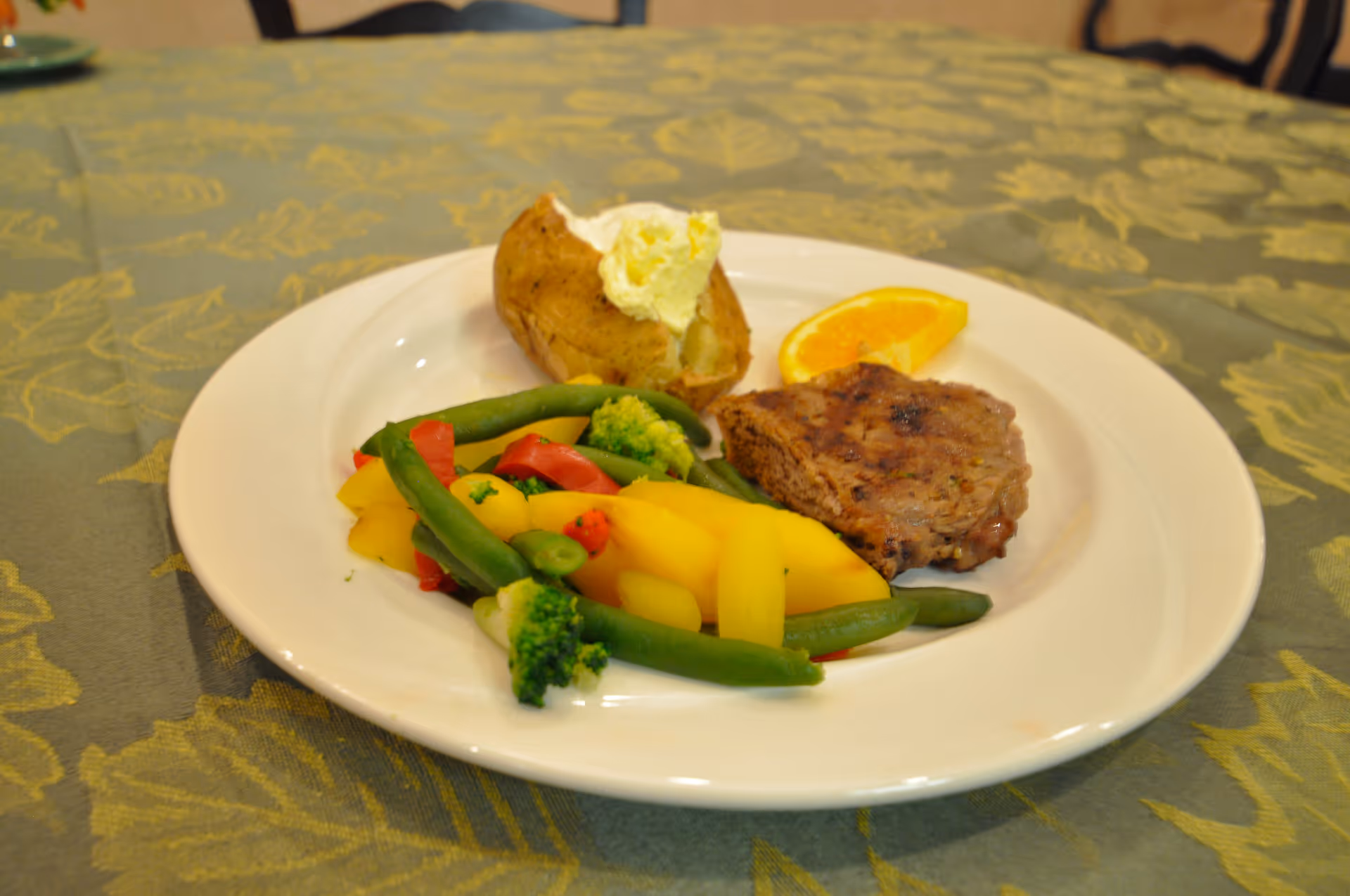 A white plate with a baked potato topped with butter, a slice of orange, a piece of grilled meat, and a serving of mixed vegetables including green beans, broccoli, red bell peppers, and yellow squash on a green patterned tablecloth.