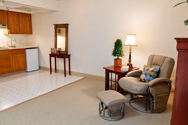 A cozy interior space featuring a small kitchen area with wooden cabinets, a sink, and a mini refrigerator on the left. Adjacent to the kitchen is a small wooden table with a decorative mirror above it. On the right side, there is a comfortable beige recliner chair with a colorful patterned pillow, accompanied by a matching footrest. Next to the chair is a wooden side table with a small potted plant, a table lamp, and some books. The floor transitions from a tiled kitchen area to carpeted living space.