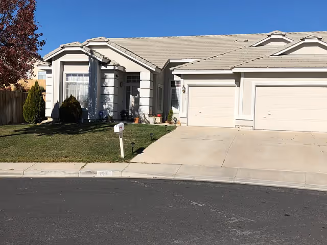 Front exterior view of a single-story residential house with a light-colored roof and walls, a two-car garage, a small lawn with a mailbox, and a clear blue sky.
