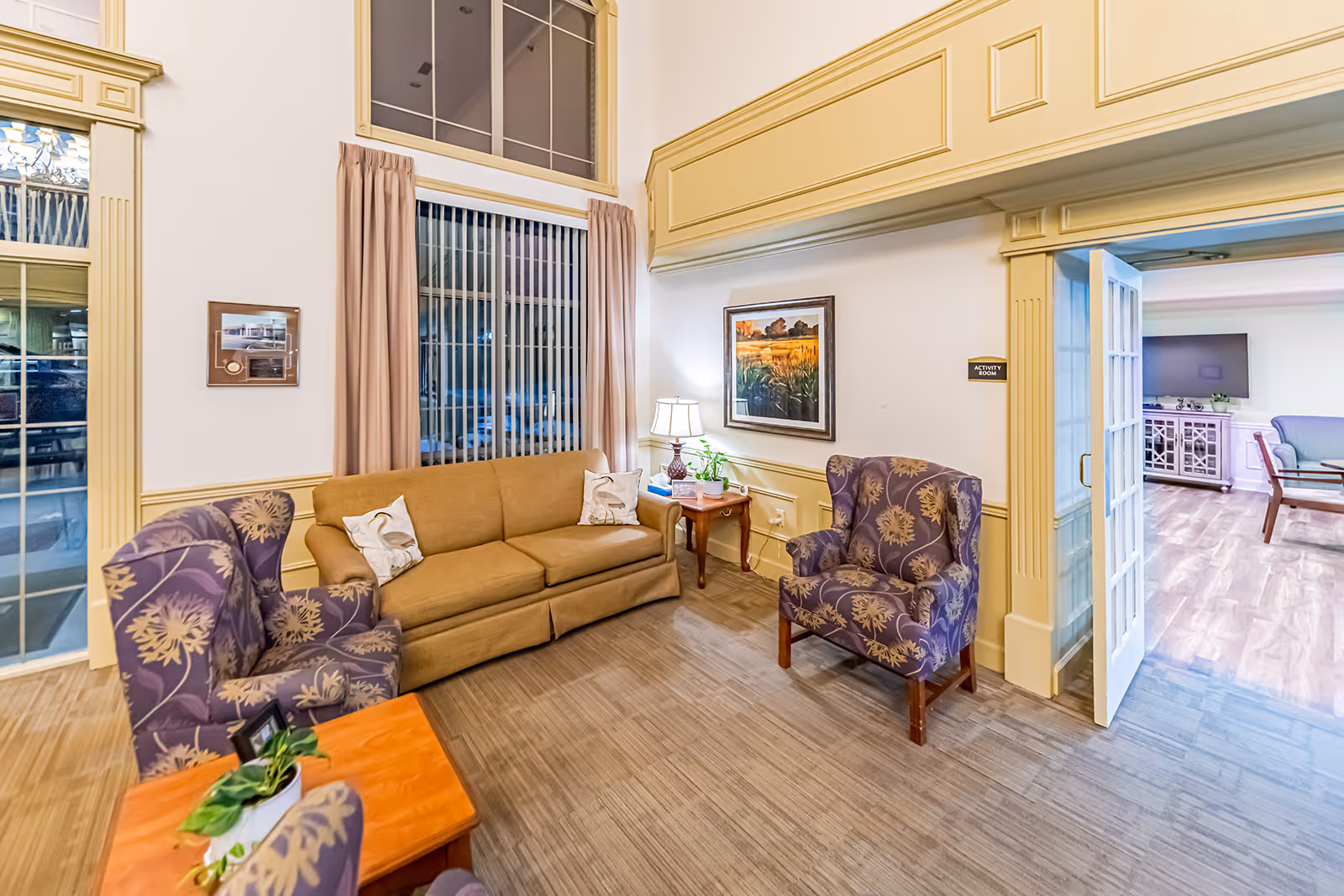 A cozy seating area in an independent living facility featuring a tan sofa with two decorative pillows, two purple armchairs with floral patterns, a wooden side table with a lamp and plant, and a framed landscape painting on the wall. The room has large windows with vertical blinds and beige curtains, light wood flooring, and cream-colored trim and molding. An open doorway leads to another room with additional seating and a television.