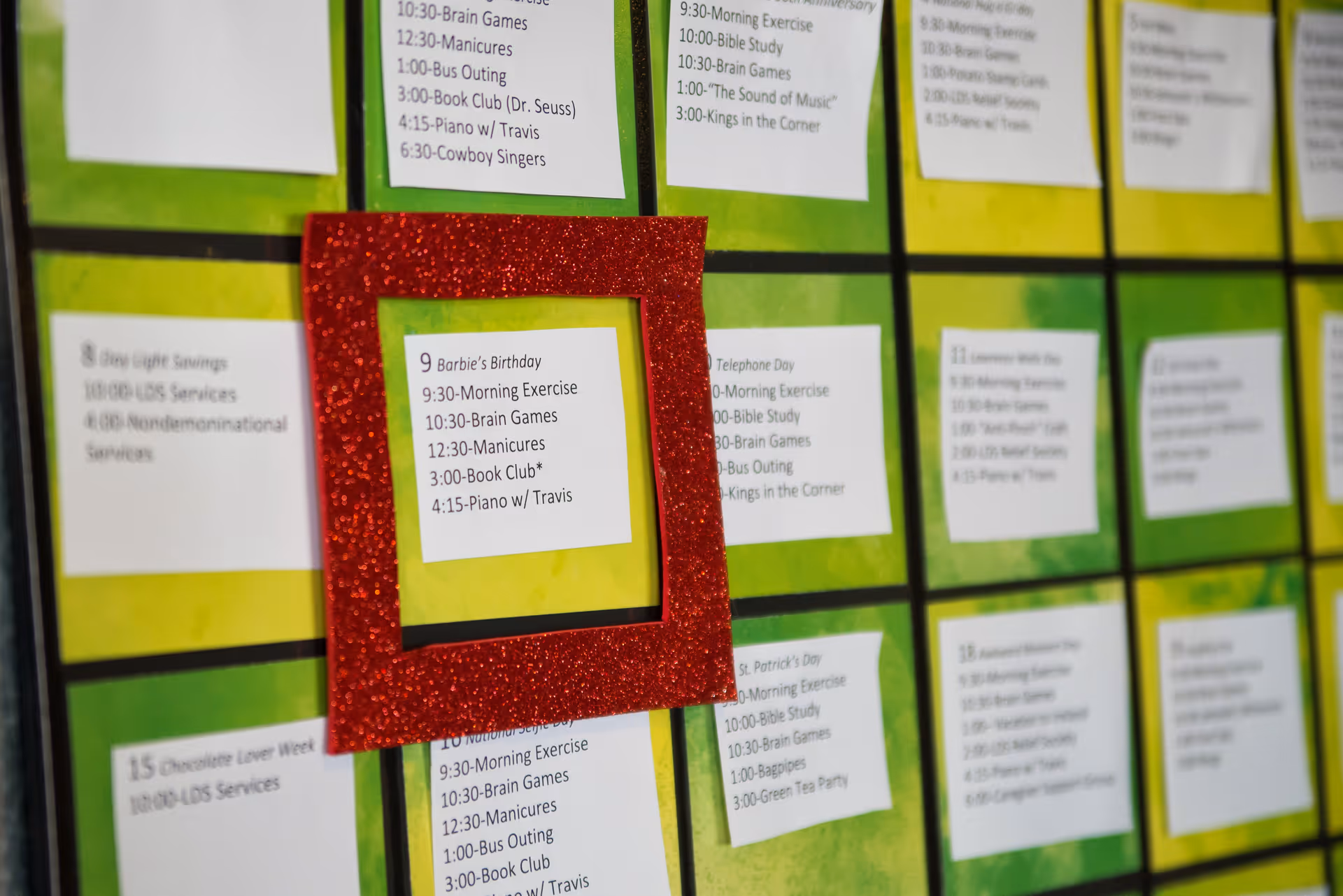 A close-up view of a colorful activity calendar with daily schedules printed on white paper squares, each placed in a grid on a green background. One day is highlighted with a red glittery frame, showing activities such as morning exercise, brain games, manicures, book club, and piano with Travis.