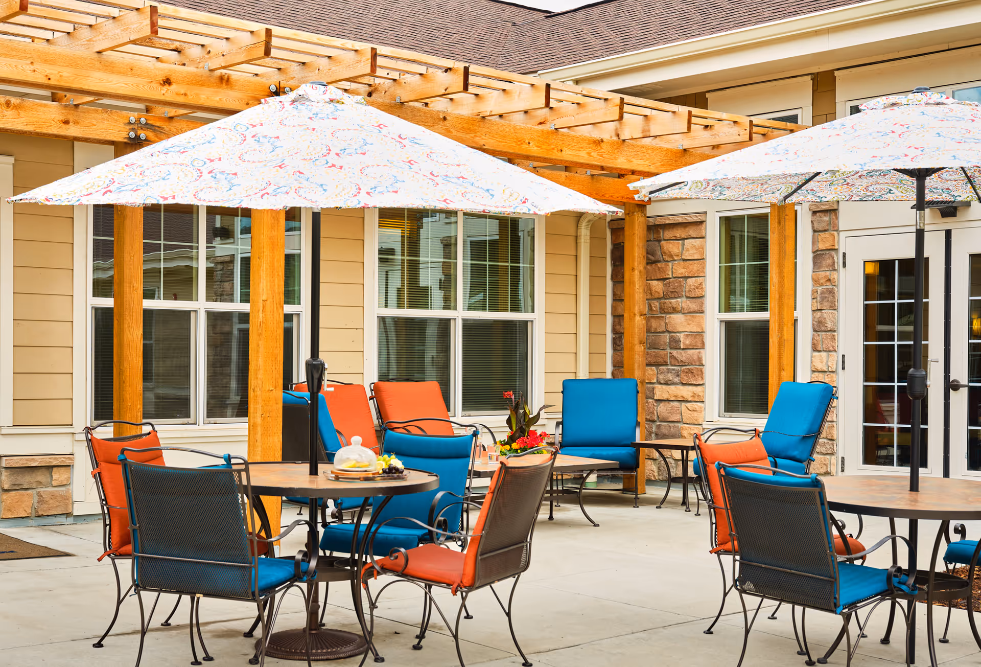 Outdoor patio area at MorningStar Assisted Living & Memory Care of Boulder with round tables, colorful cushioned chairs in blue and orange, and large patterned umbrellas providing shade. The patio is adjacent to a building with beige siding, stone accents, and windows.