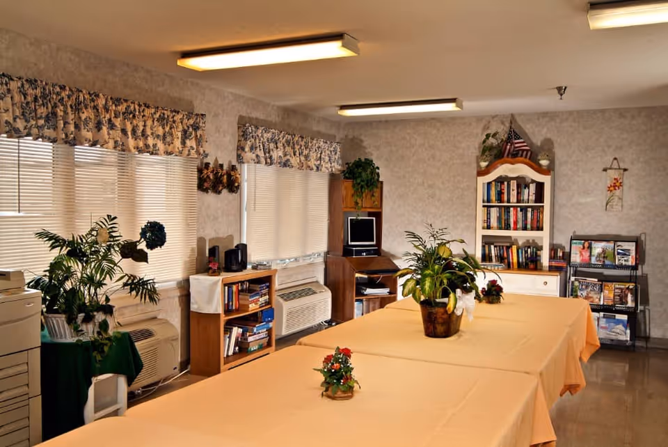 A cozy common area with two tables covered in beige tablecloths, decorated with potted plants and small flower arrangements. The room has several windows with floral valances and blinds, bookshelves filled with books, a small computer desk with a monitor, and a magazine rack. The walls are light-colored, and the ceiling has fluorescent lights.