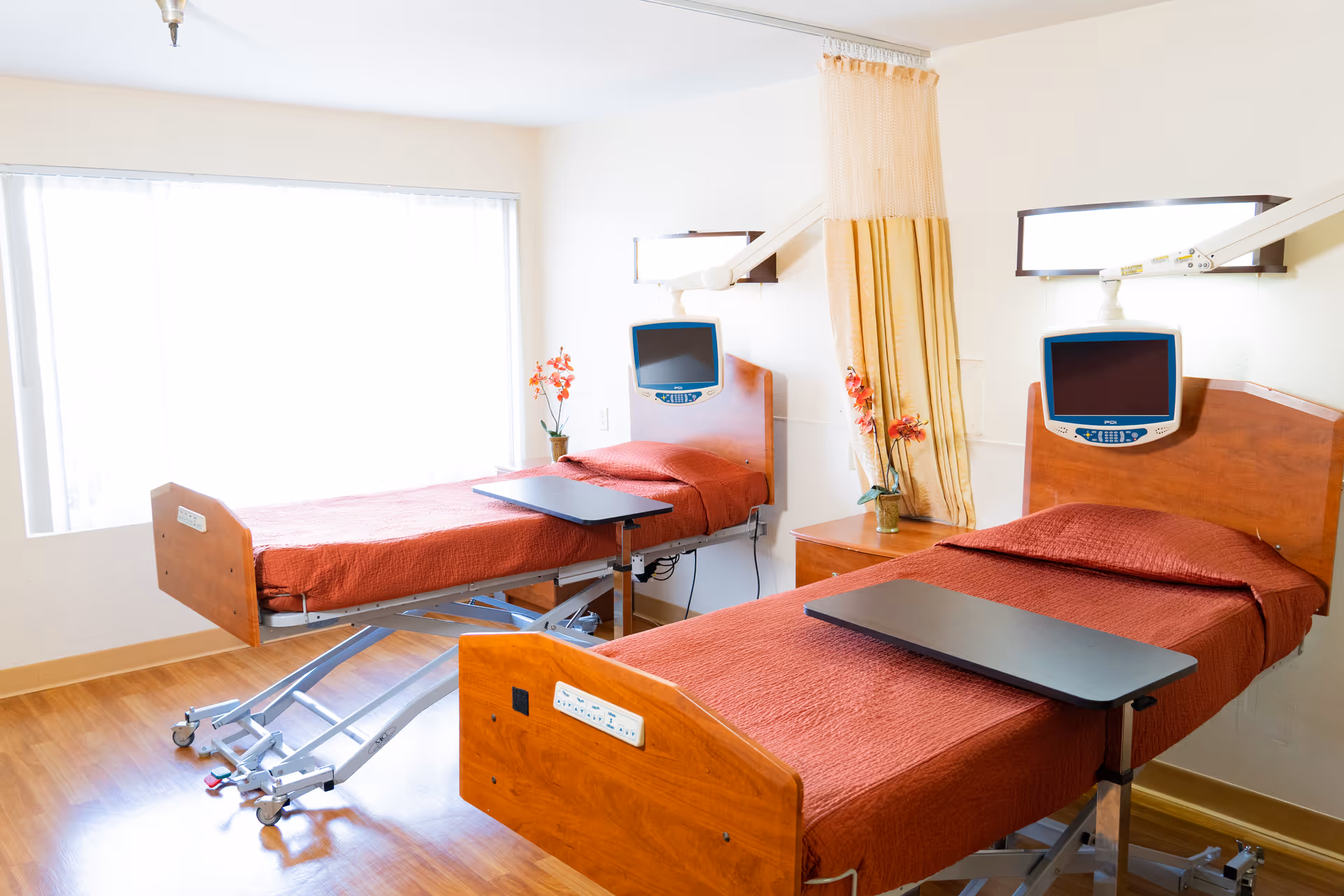 A bright room with two hospital-style beds covered in red bedspreads, each equipped with an adjustable overbed table and a mounted television screen above the headboard. There is a window with sheer curtains letting in natural light, a small nightstand with a vase of flowers, and a privacy curtain between the beds.