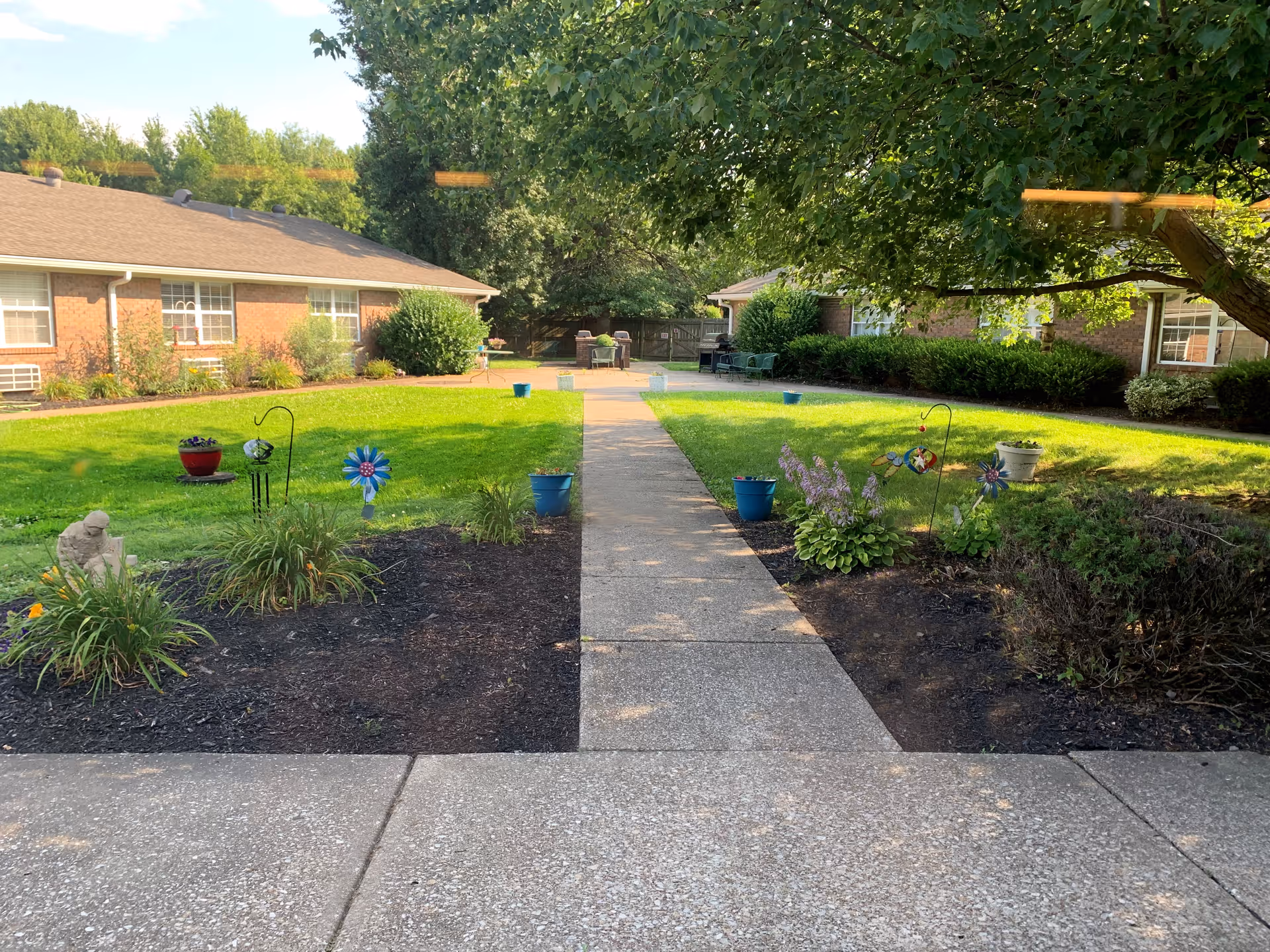 A sunny outdoor courtyard area with a concrete walkway leading through a green lawn and flower beds. There are small decorative garden stakes and potted plants along the path. Two single-story brick buildings with windows and bushes surround the courtyard. A large tree provides shade on the right side.