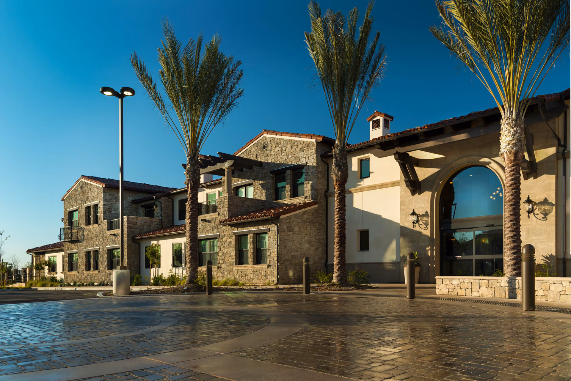Stone-clad senior living building front with palm trees, arched entrance and a paved driveway under a clear blue sky.