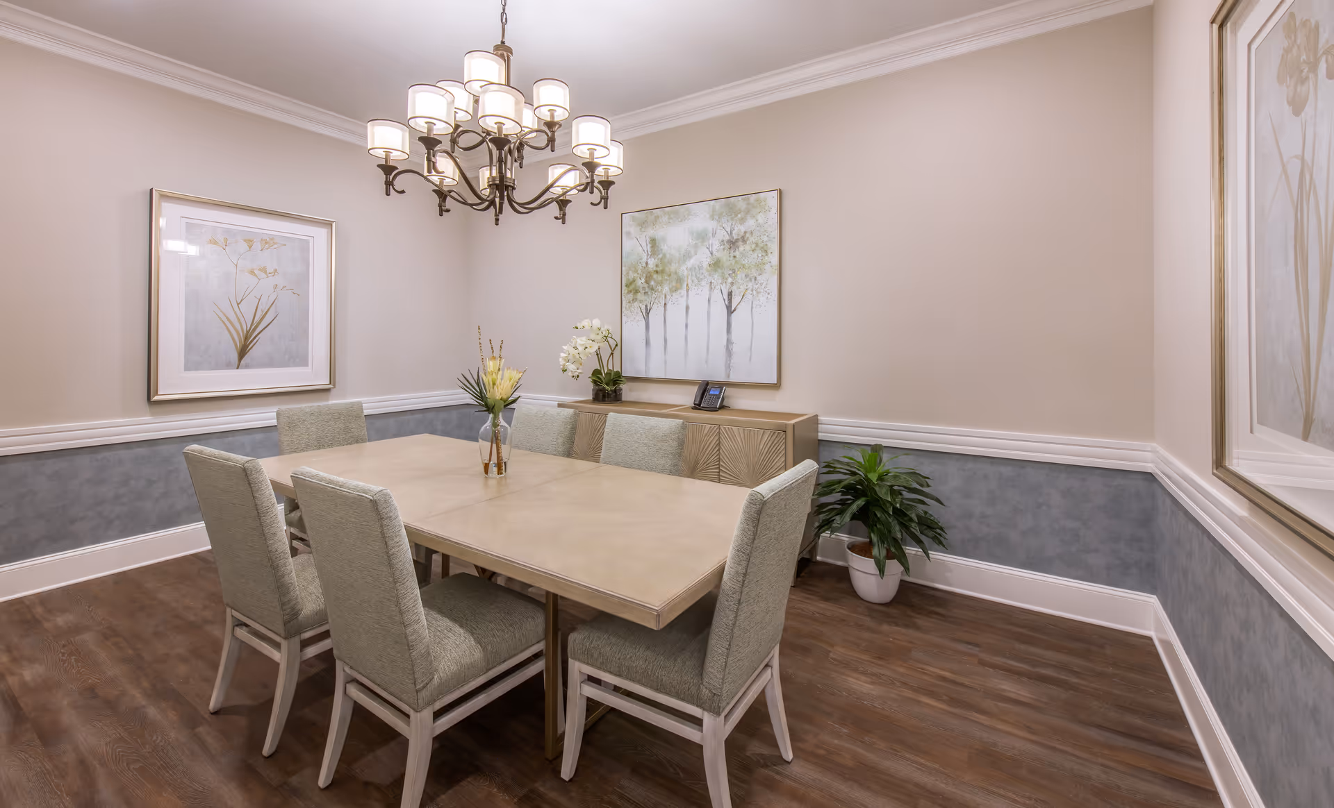 A dining room with a rectangular wooden table surrounded by six upholstered chairs. The room features a chandelier with multiple lights hanging above the table, two framed botanical paintings on the walls, a sideboard with a telephone and decorative plants, and a potted plant on the floor. The walls are painted beige with a blue-gray wainscoting, and the floor is wood.