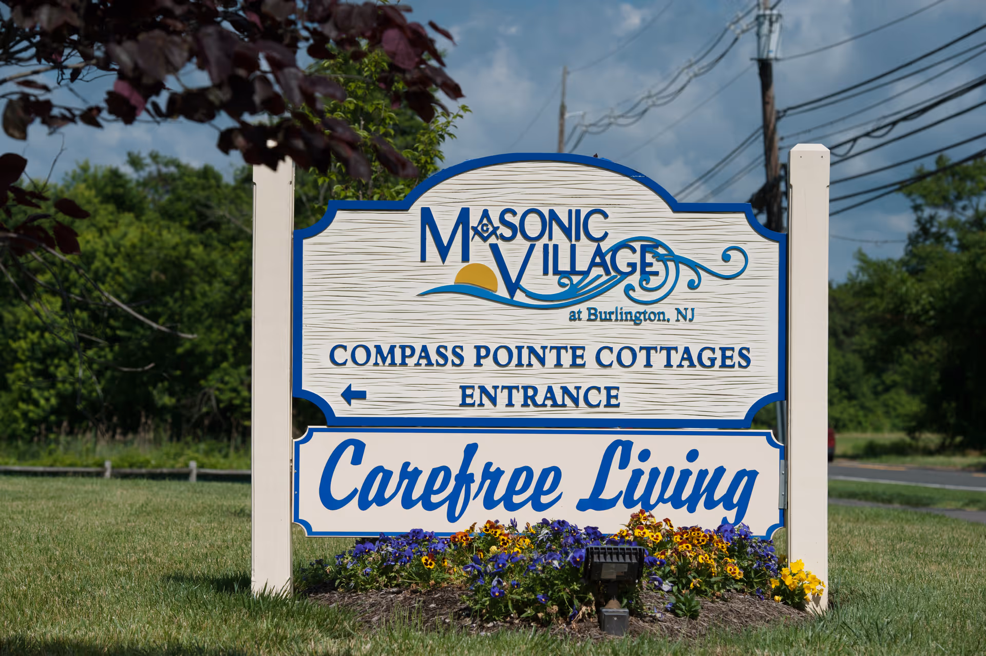 Outdoor sign for Masonic Village at Burlington, NJ, indicating the entrance to Compass Pointe Cottages with the phrase 'Carefree Living' below. The sign is surrounded by grass and flowers, with trees and power lines in the background.