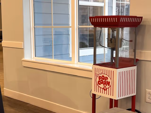 A red and white vintage-style popcorn machine stands near a window inside a room with beige walls and wooden flooring. The popcorn machine has the words 'Fresh Pop Corn' written on its front panel.