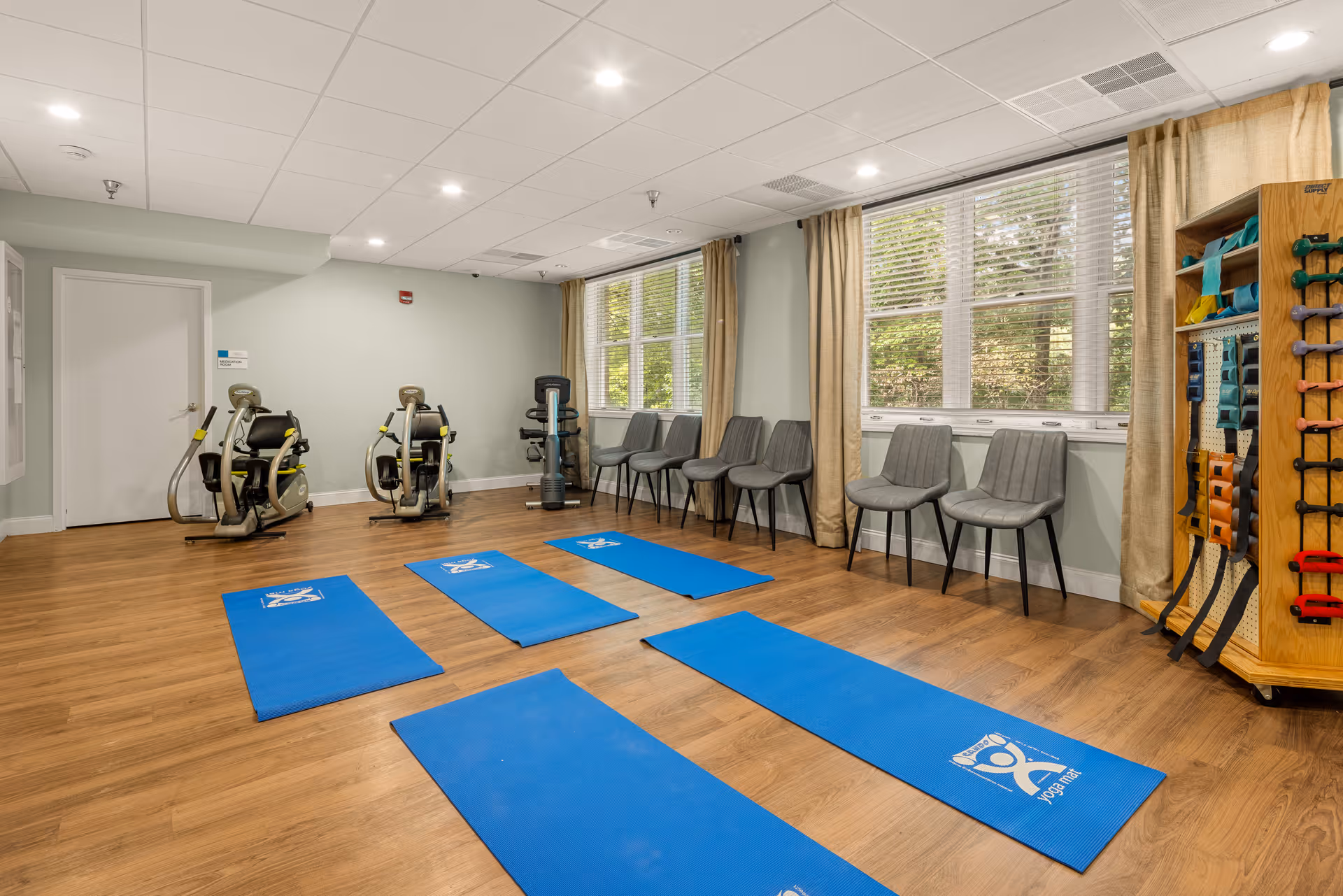 A senior living facility exercise room with wooden flooring, five blue yoga mats laid out on the floor, two exercise machines against the far wall, a row of gray chairs along the windowed wall, and a rack holding various exercise bands and weights.