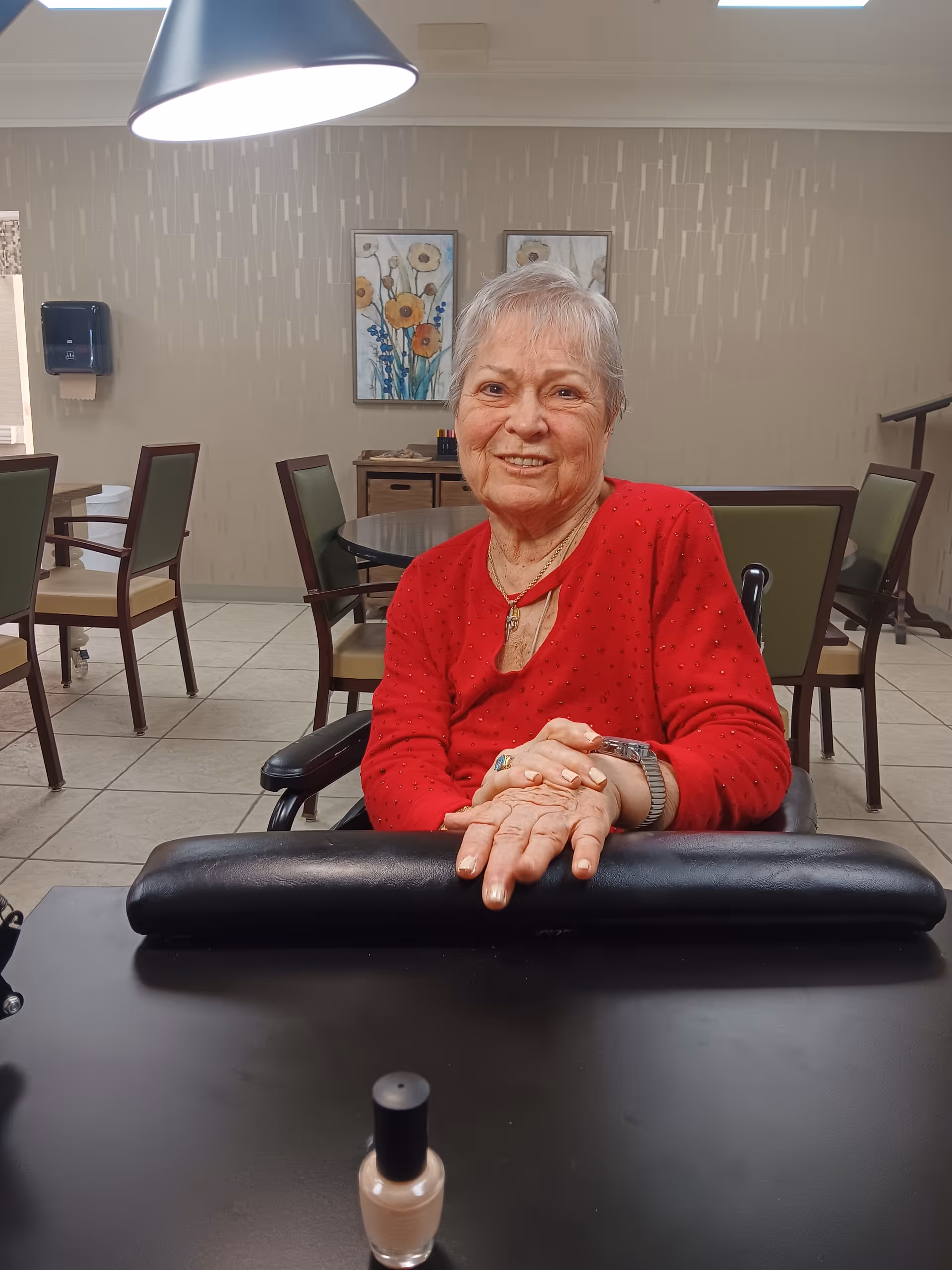 An elderly woman with short gray hair wearing a red sweater is sitting at a table in a room with beige walls and floral paintings. She is smiling and resting her hands on a black armrest on the table. A bottle of light-colored nail polish is on the table in front of her. The room has several chairs and tables, and a paper towel dispenser is mounted on the wall.