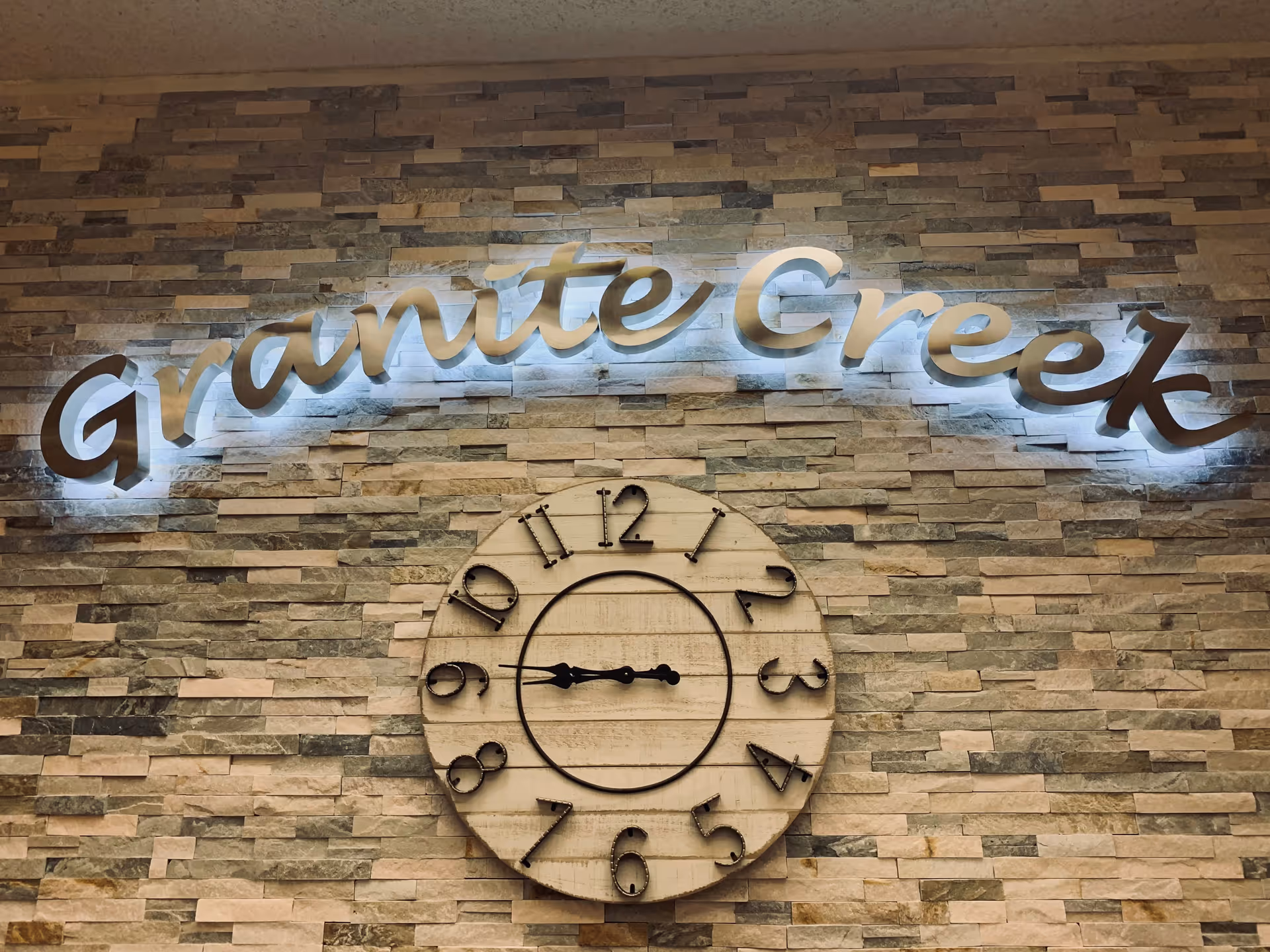 An illuminated "Granite Creek" script sign and a large rustic round clock mounted on a stacked stone interior wall.