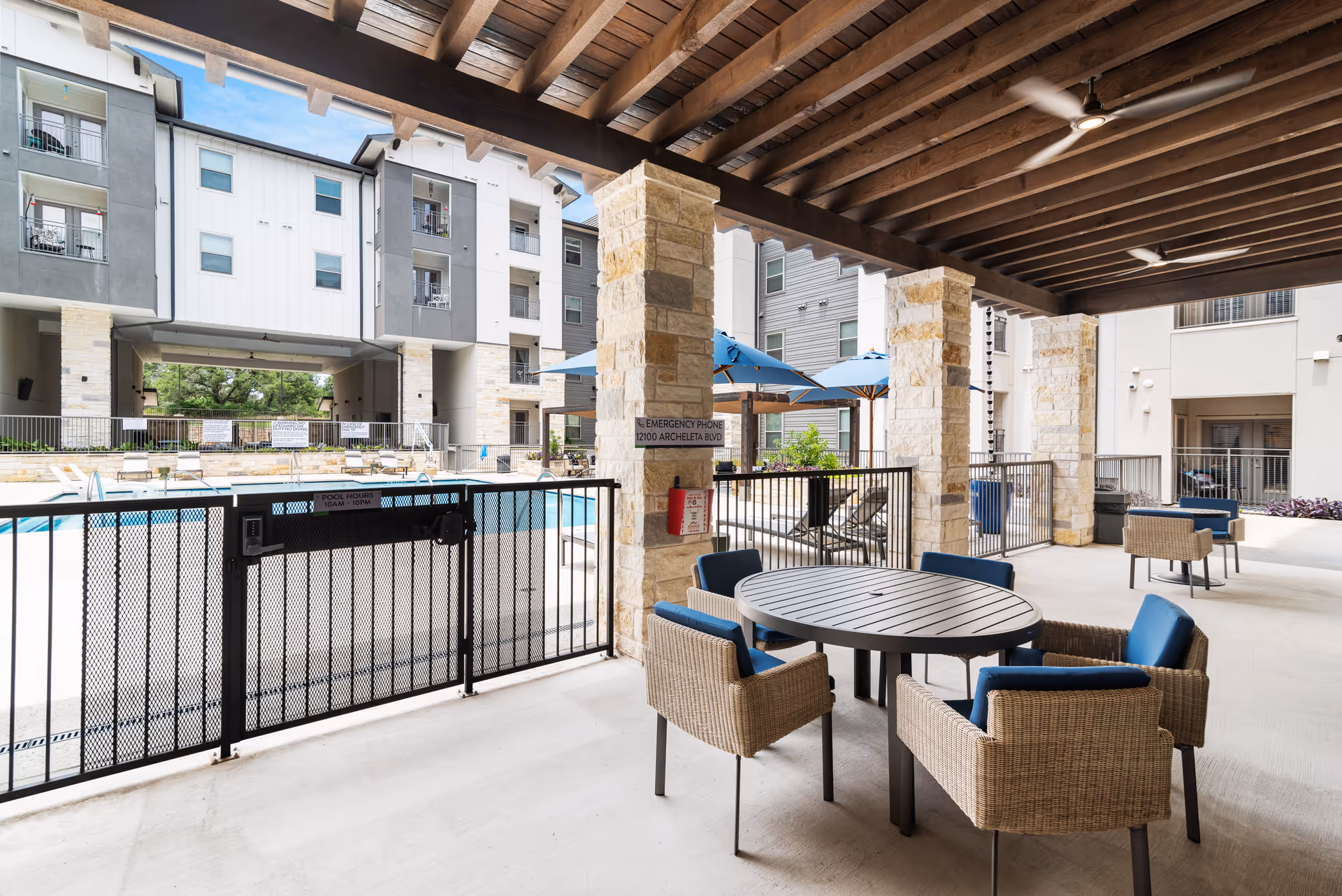 Covered outdoor seating area with round tables and wicker chairs with blue cushions, overlooking a fenced swimming pool with lounge chairs and blue umbrellas. The area is part of a multi-story residential building with balconies and stone pillars supporting a wooden pergola ceiling.