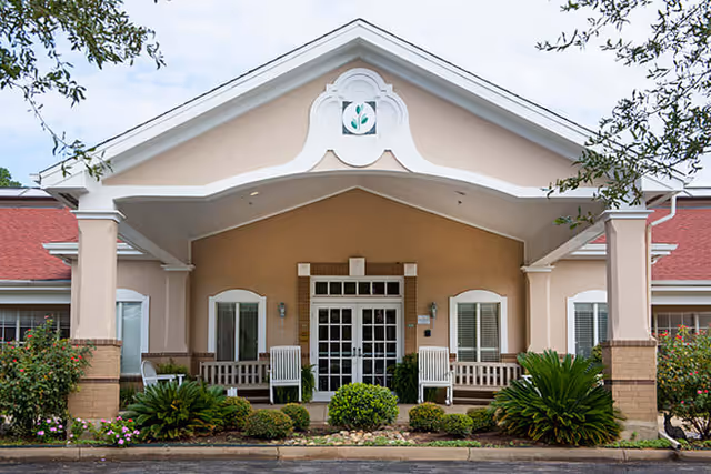Front exterior view of a single-story building with a covered entrance, white framed glass doors, two white rocking chairs on the porch, and well-maintained shrubs and plants in front.