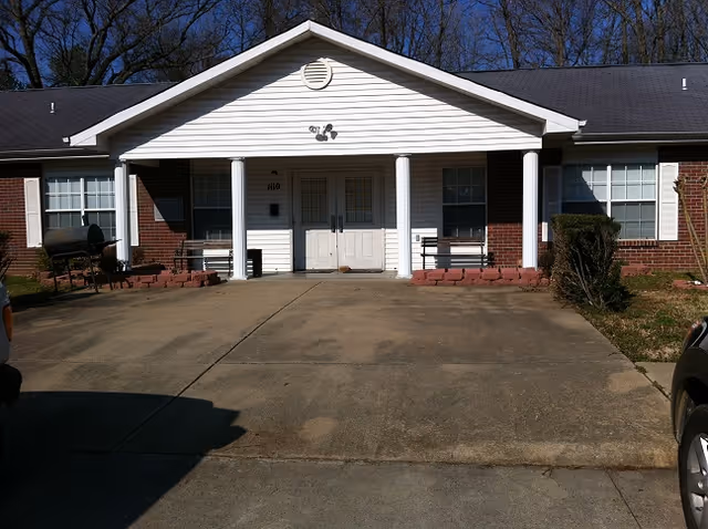 Front exterior view of a single-story brick and white siding building with a covered entrance supported by two white columns. There are two benches on either side of the entrance door, windows with white blinds, and a concrete driveway leading up to the entrance. Trees are visible in the background under a clear blue sky.