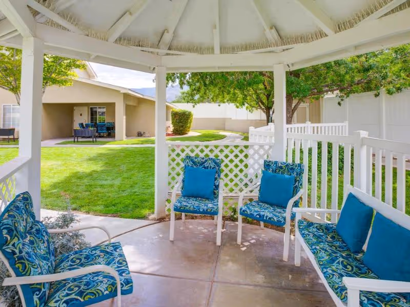View from inside a white gazebo with blue cushioned chairs and benches, overlooking a green lawn with trees and a beige building in the background under a partly cloudy sky.