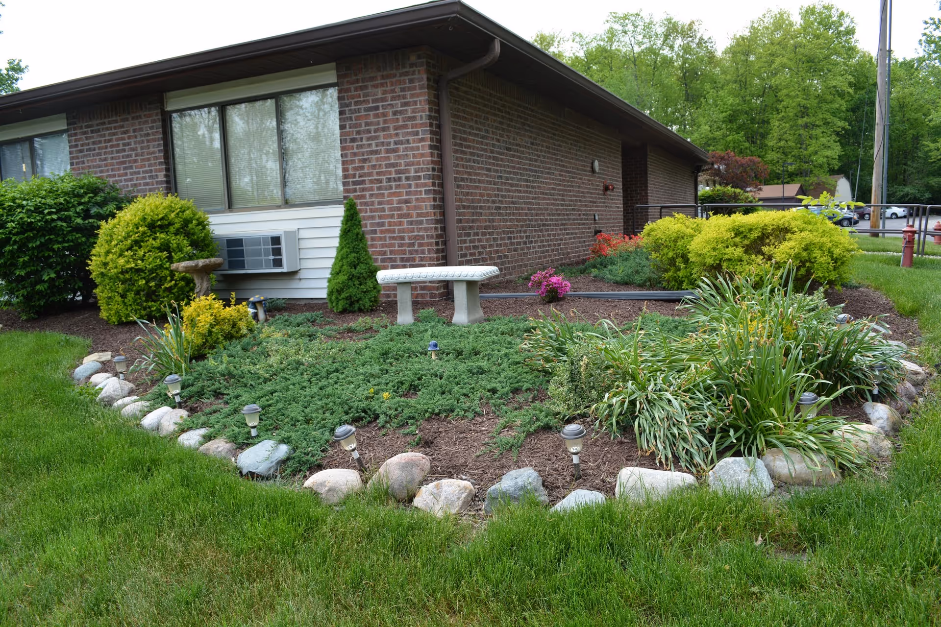 A landscaped garden area outside a brick building with various green shrubs, plants, and a small white stone bench. The garden is bordered by rocks and has several small solar lights. There is a window with an air conditioning unit on the building wall.