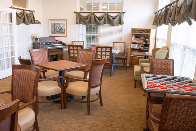 A cozy common room with several tables and chairs arranged for group activities. There is a checkers game set up on one table, a small organ against the wall, and a wooden shelving unit with books and games. The room has large windows with green valance curtains letting in natural light.