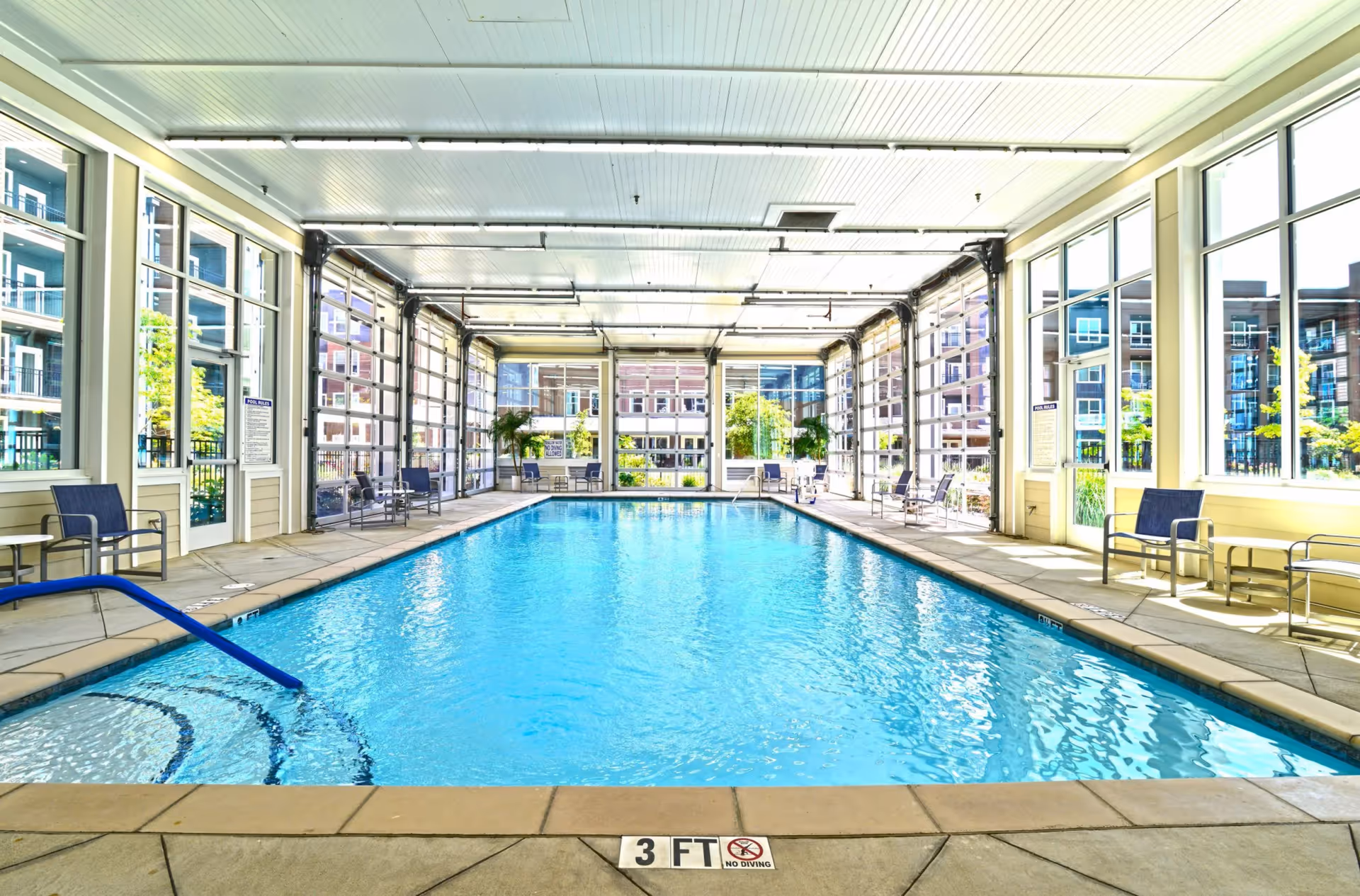 Indoor swimming pool with large windows and seating along both sides in a bright amenity room.