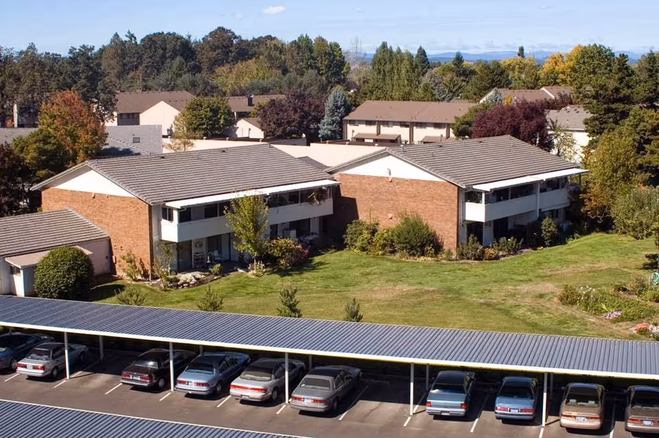 Aerial view of Friendsview Retirement Community showing two brick residential buildings with balconies, surrounded by green lawns and trees. In the foreground, there is a covered parking area with several parked cars. The background features more trees and residential buildings under a clear sky.
